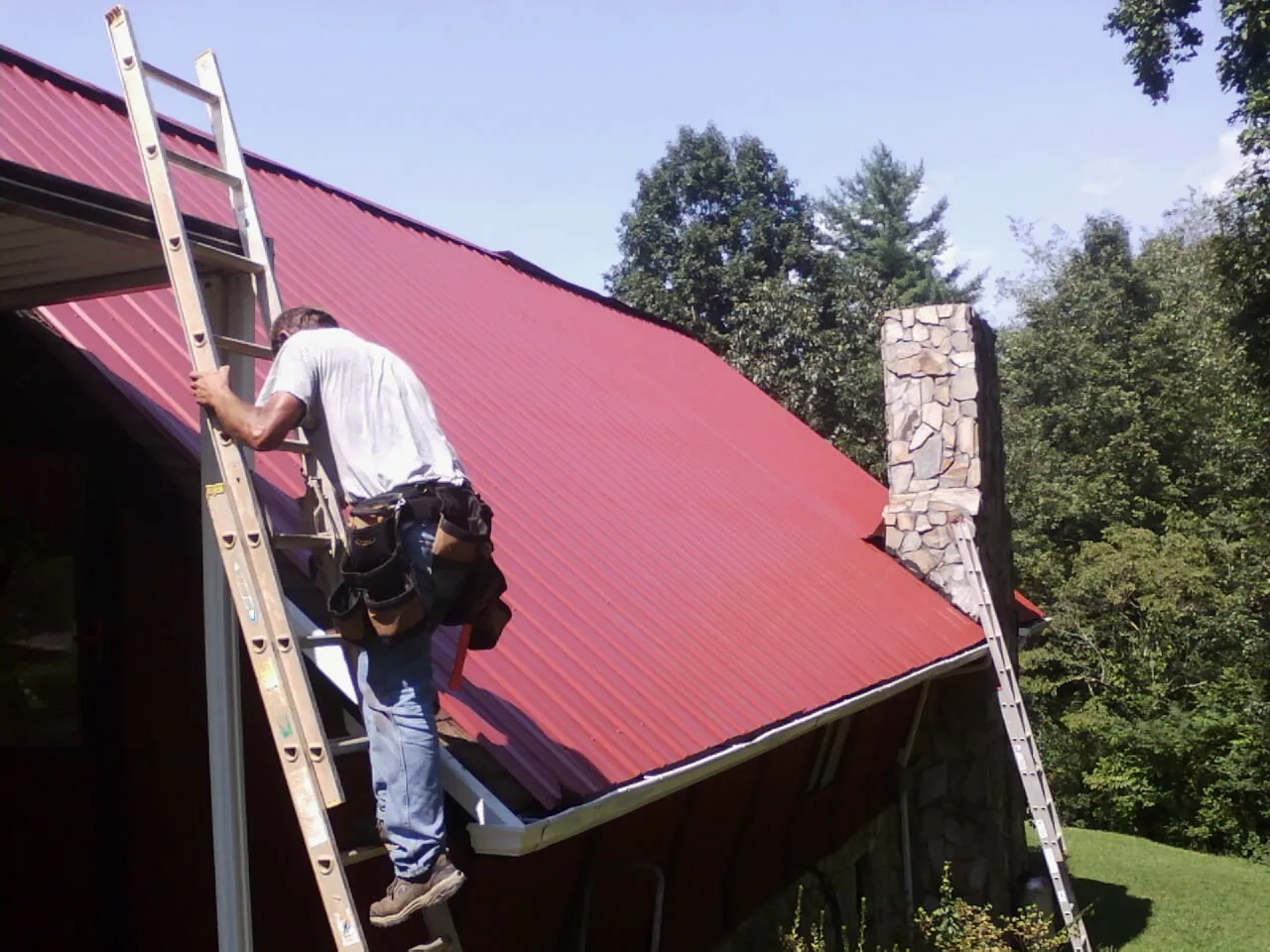 Roofer on a ladder beside a red metal roof and stone chimney — Soffit & Fascia Repair by Roofing Brothers in Mattoon